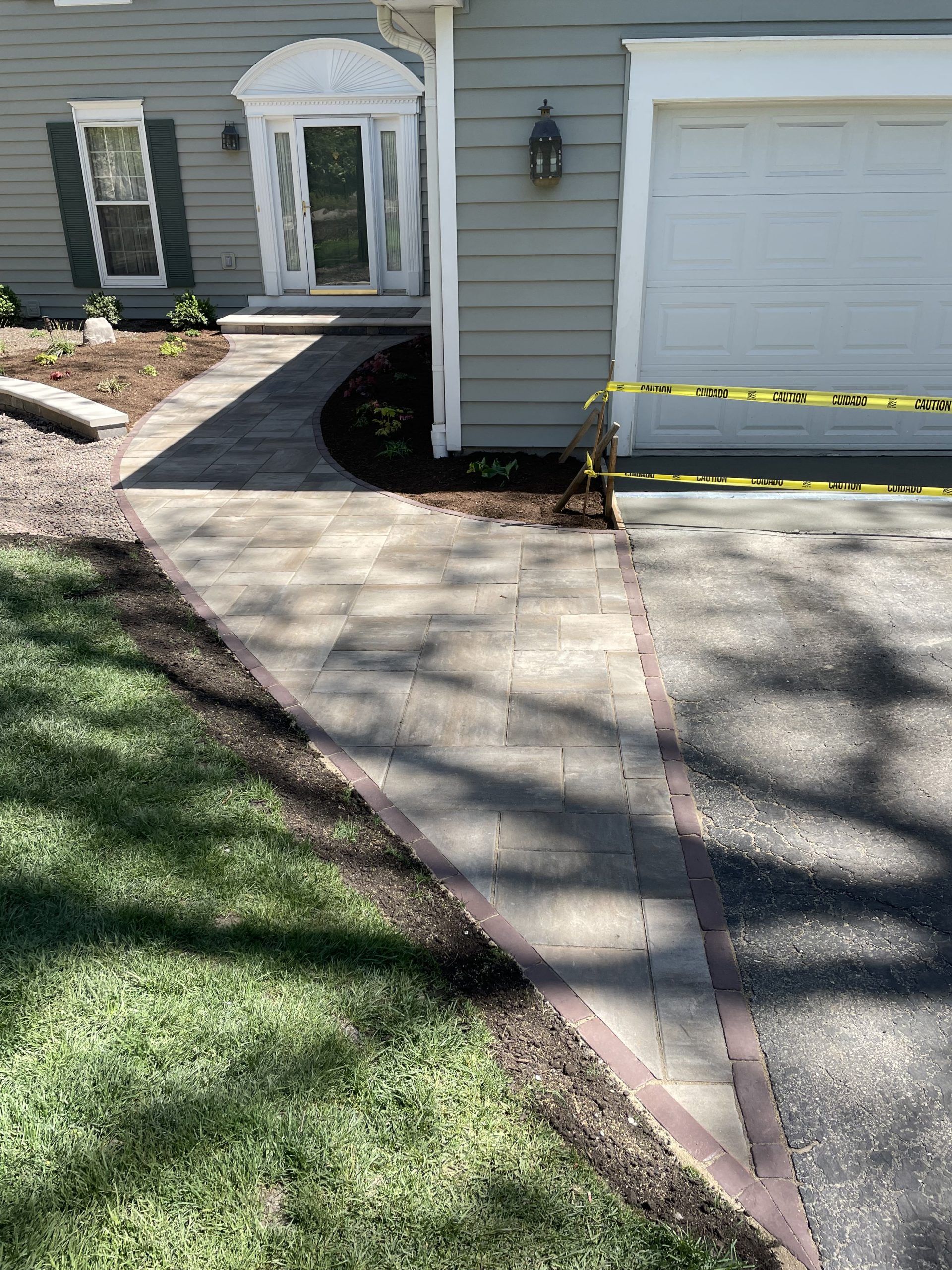 Paved walkway with brick border leads to a house entrance. Green lawn and driveway. Yellow caution tape.