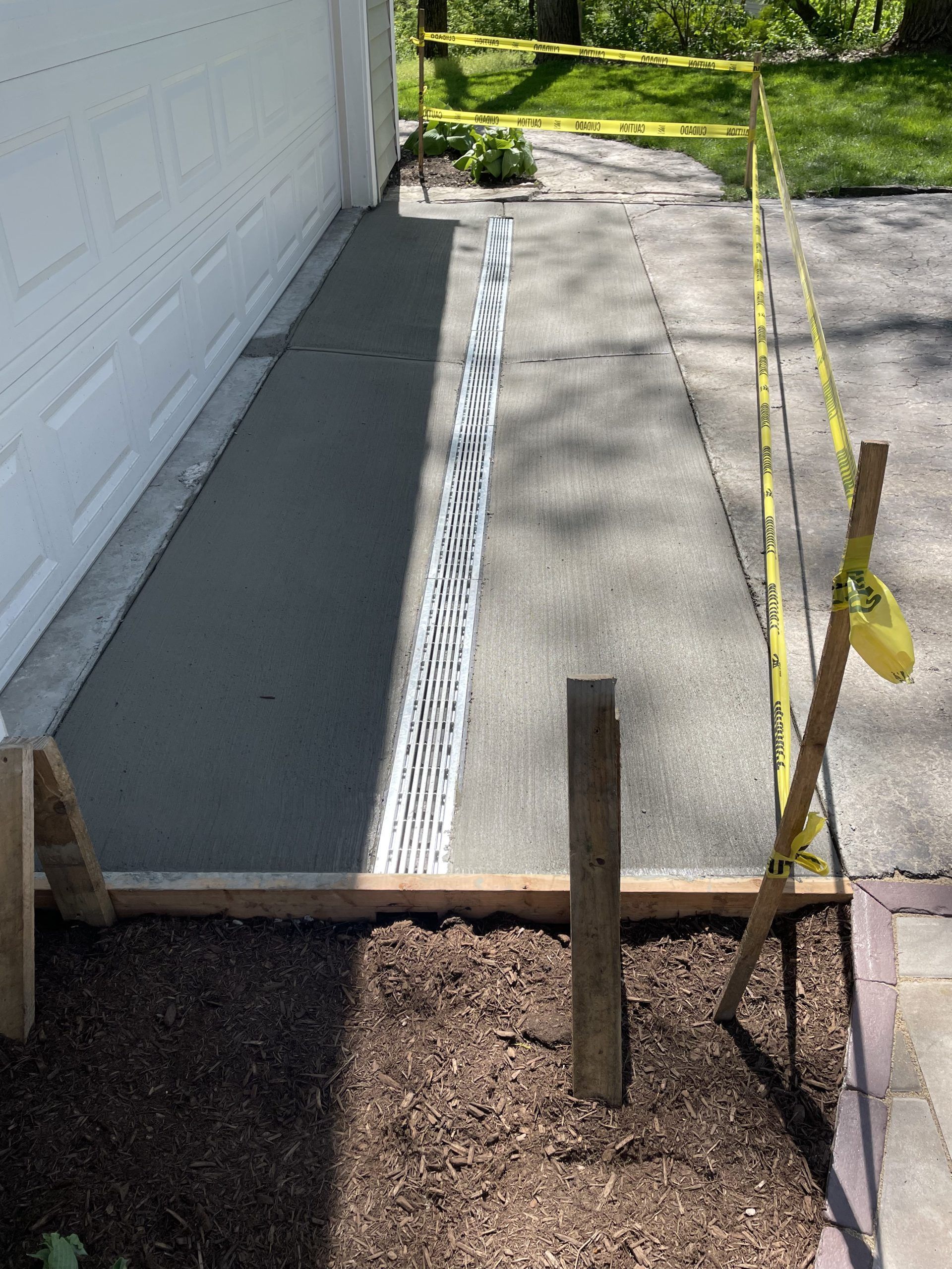 Freshly poured concrete walkway next to a white wall and a brown mulch bed, caution tape.
