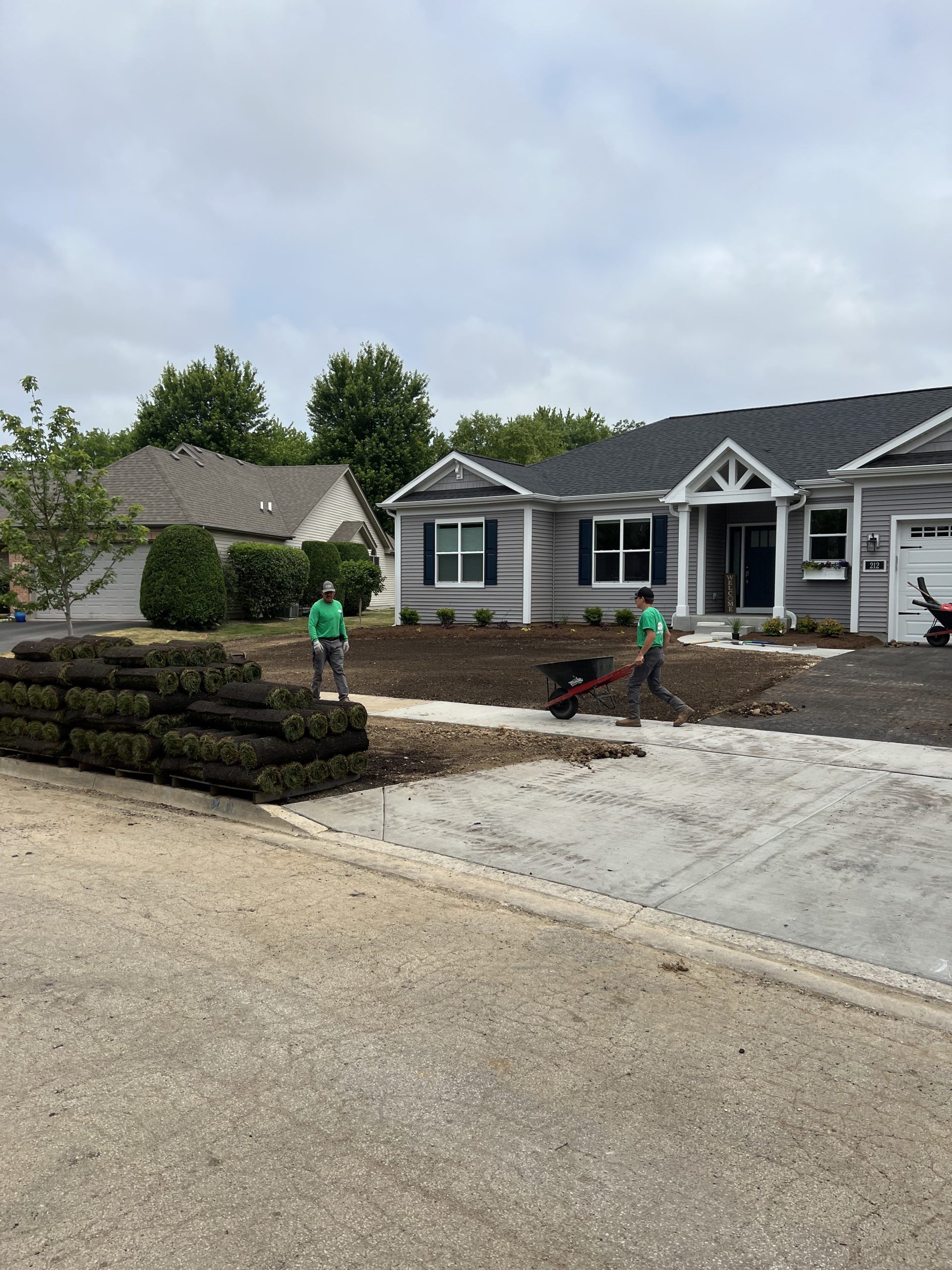 Two people laying sod in front of a gray house. Sod bricks are stacked. A wheelbarrow sits nearby.