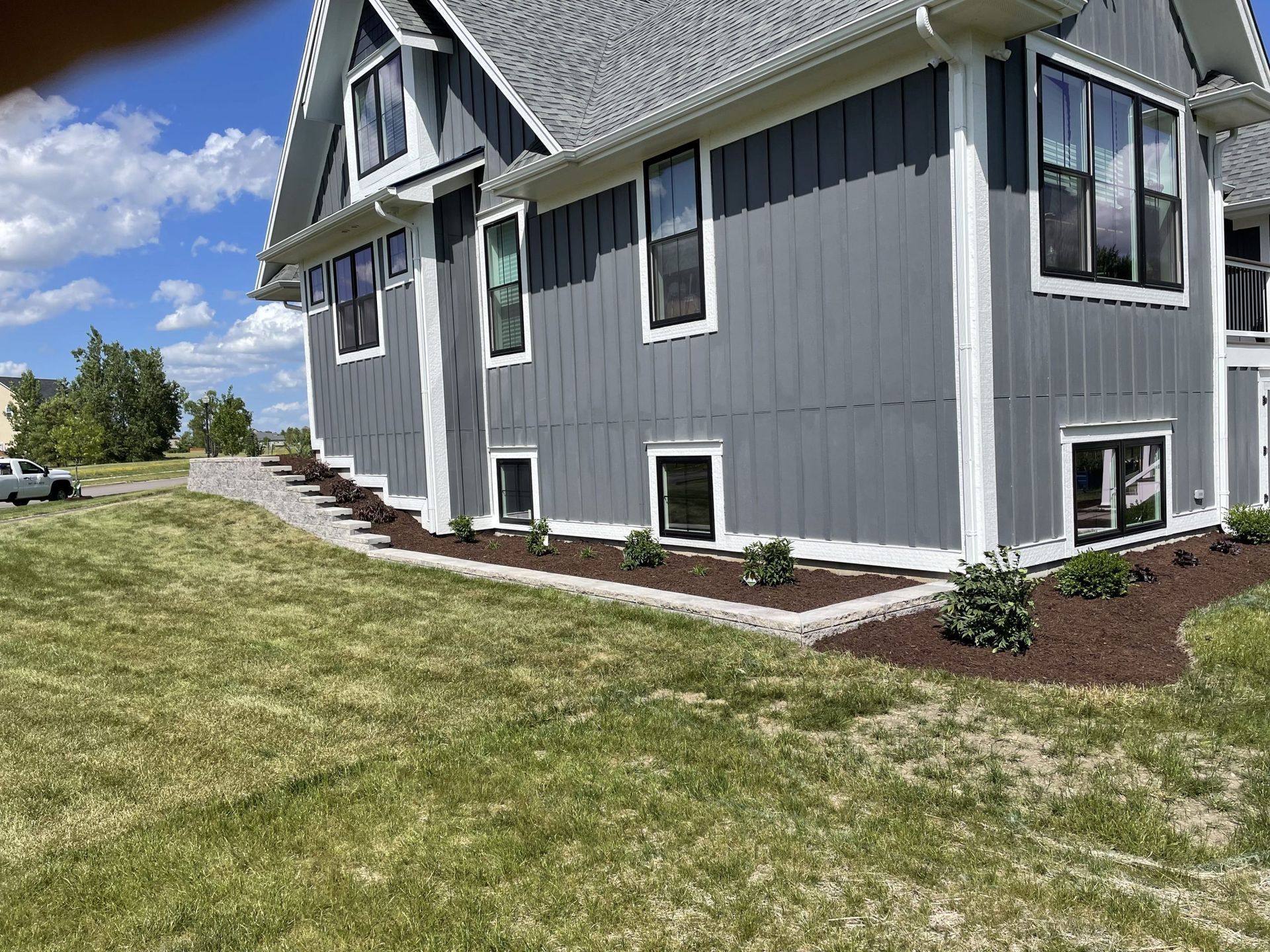Gray house with white trim, landscaped border of mulch and plants, green lawn, blue sky.
