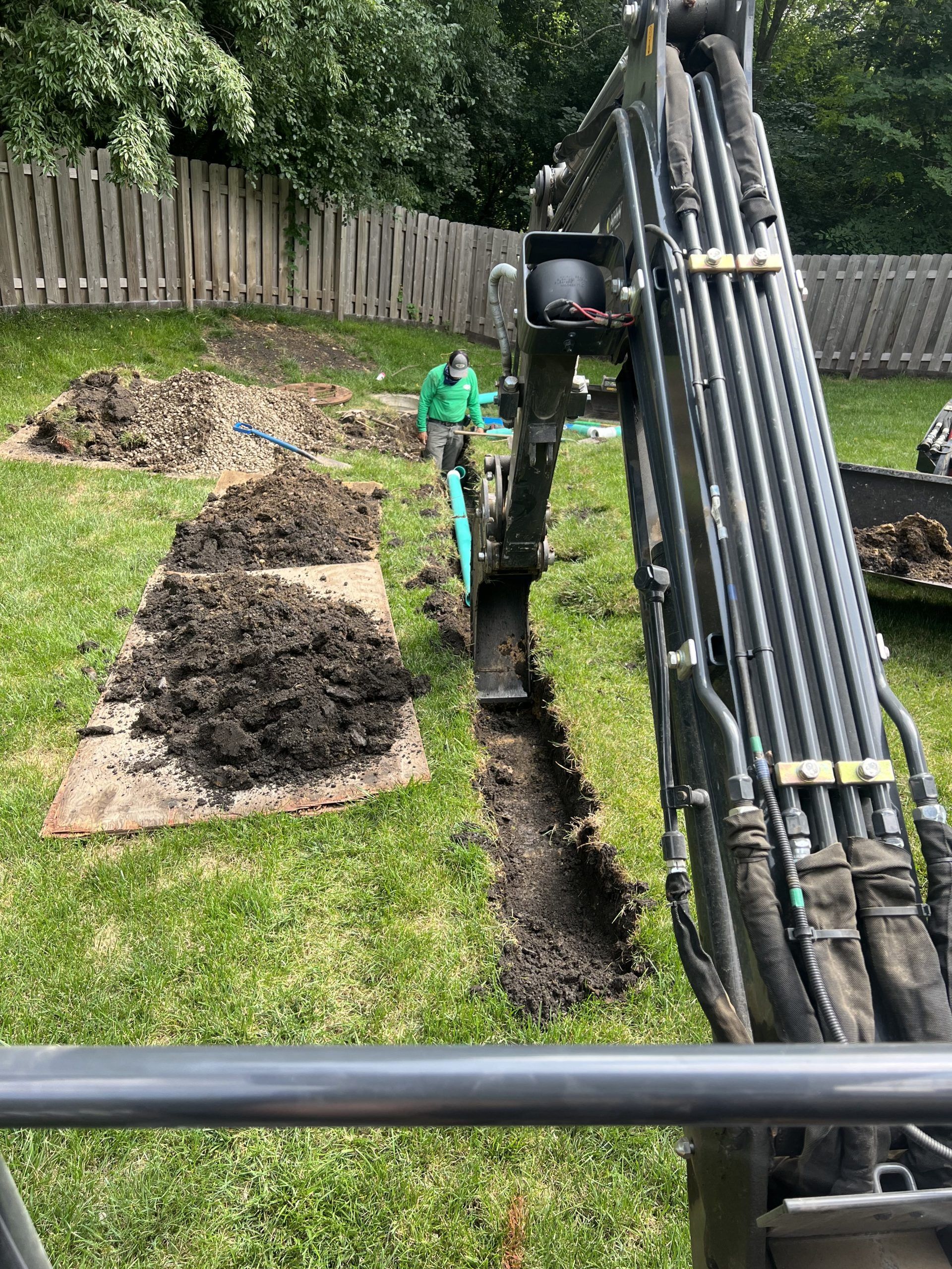 Mini excavator digging a trench in a grassy backyard, piles of dirt nearby. Man in green jacket watches.