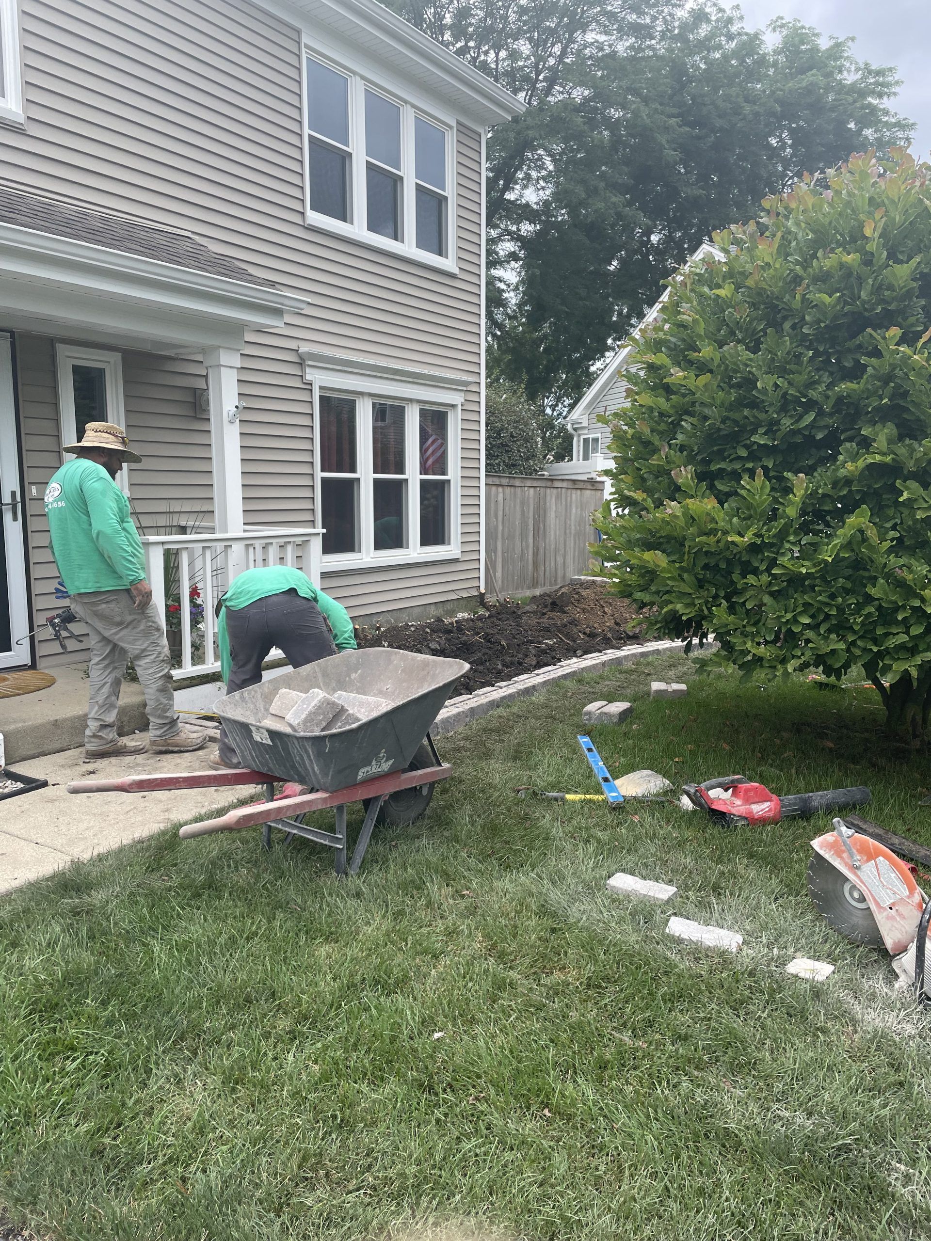 Two people working on landscaping near a two-story beige house. One is bending over a wheelbarrow. Green and gray clothing.