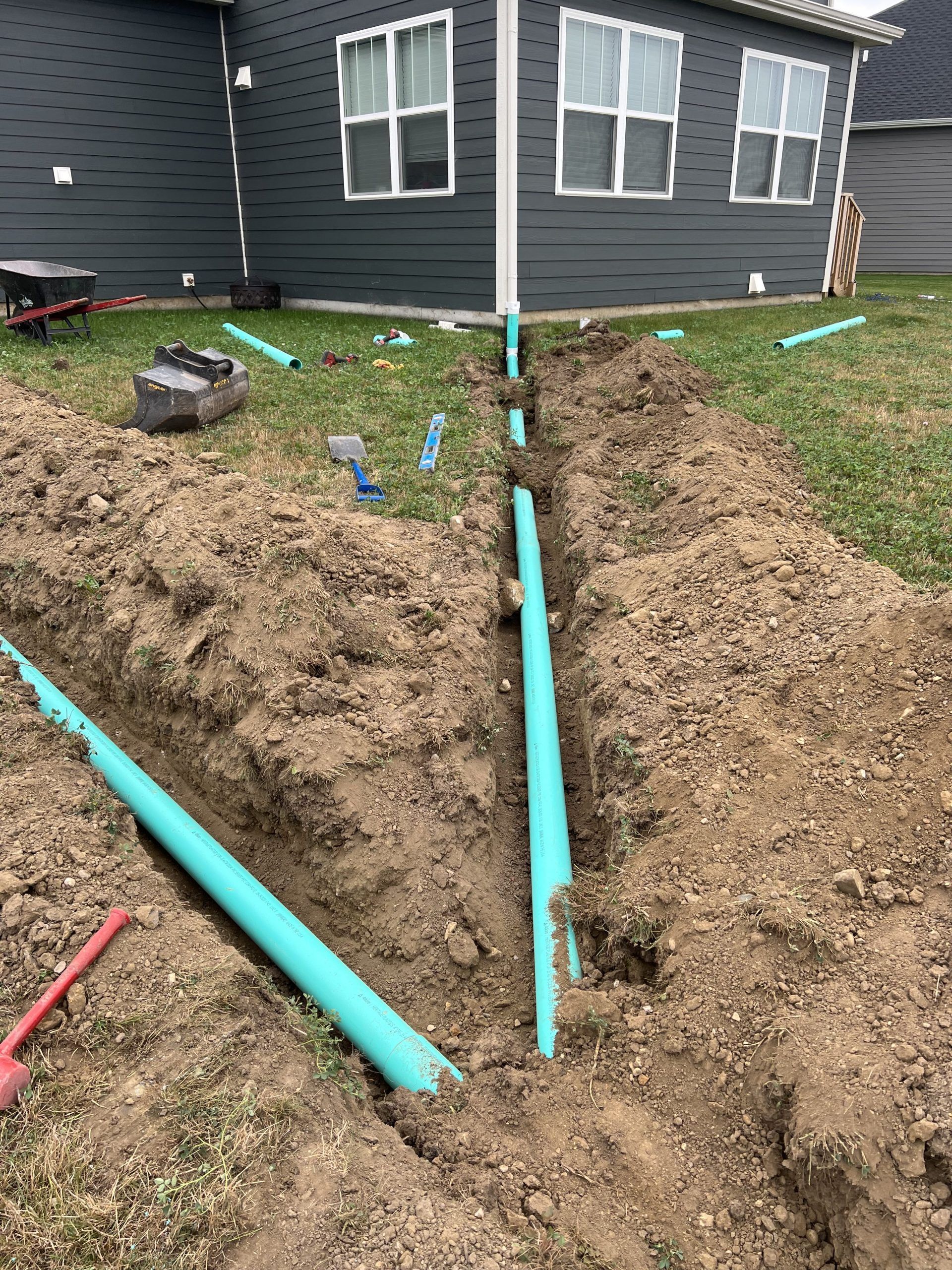 Trench with blue pipes in a yard near a house. Construction site with dirt and tools.