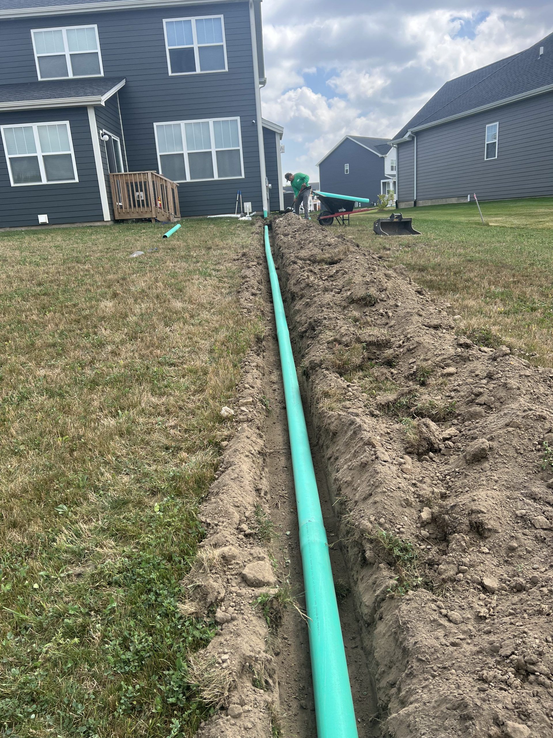 Green pipe laid in a trench in a backyard, with houses in the background.