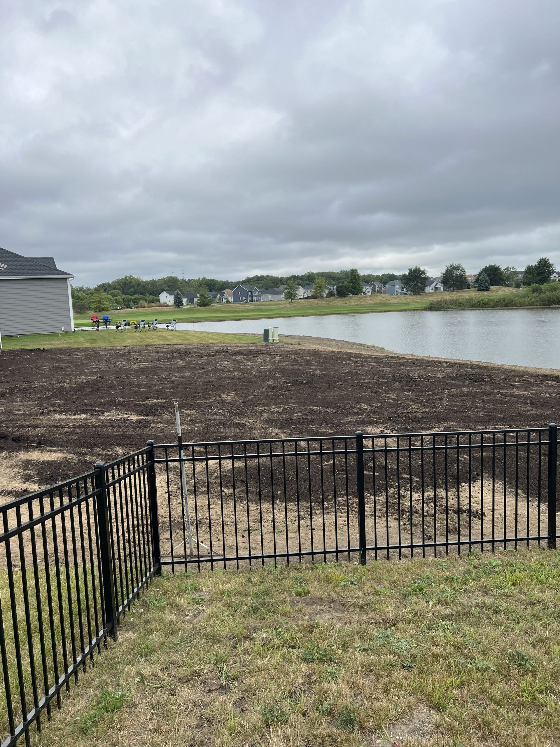 Black fence in front of a yard with dirt and a lake in the background under a cloudy sky.