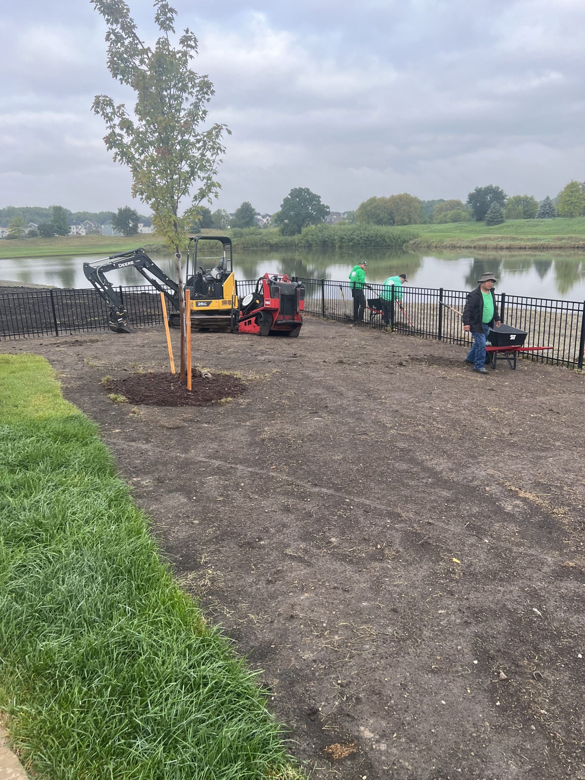 Workers planting trees and installing a border near a lake on a cloudy day.