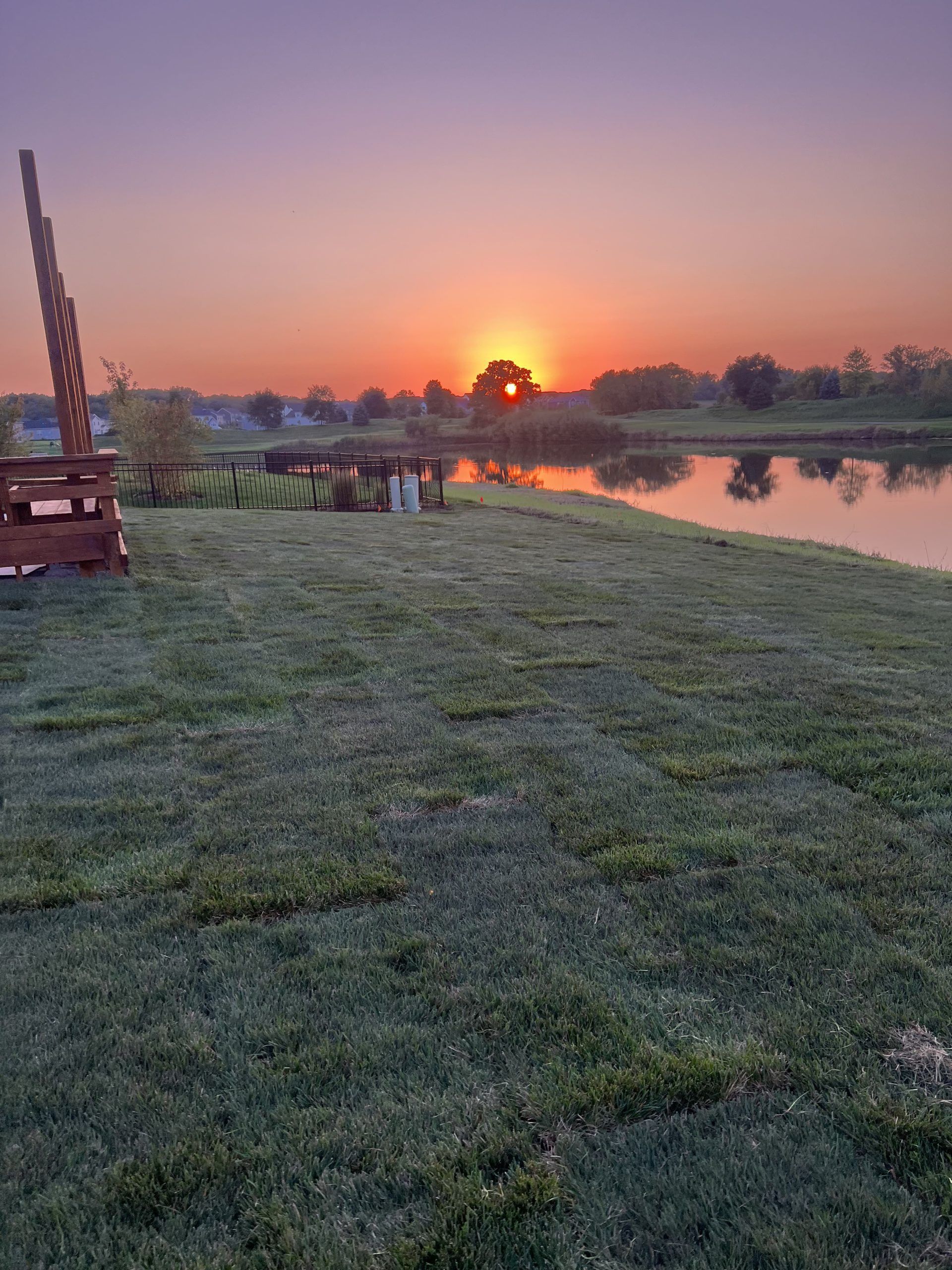 Sunrise over a pond; orange sun near treeline, reflecting in water. Green grass in foreground, wooden structure left.