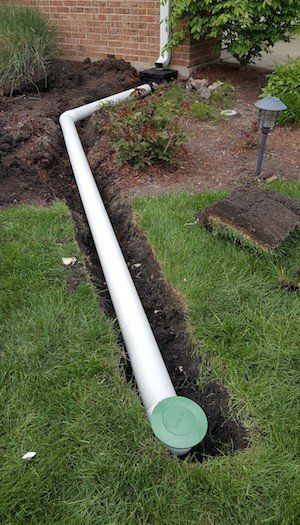 White drainage pipe in a trench, connected to a downspout, surrounded by grass and dirt.