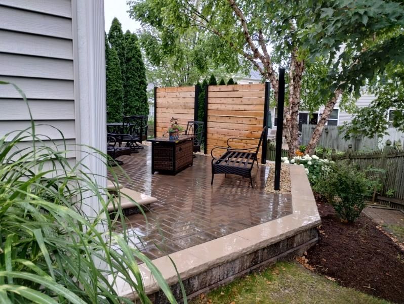 Brick patio with fire pit, seating, and wooden privacy screen in a yard with a tree and landscaping.