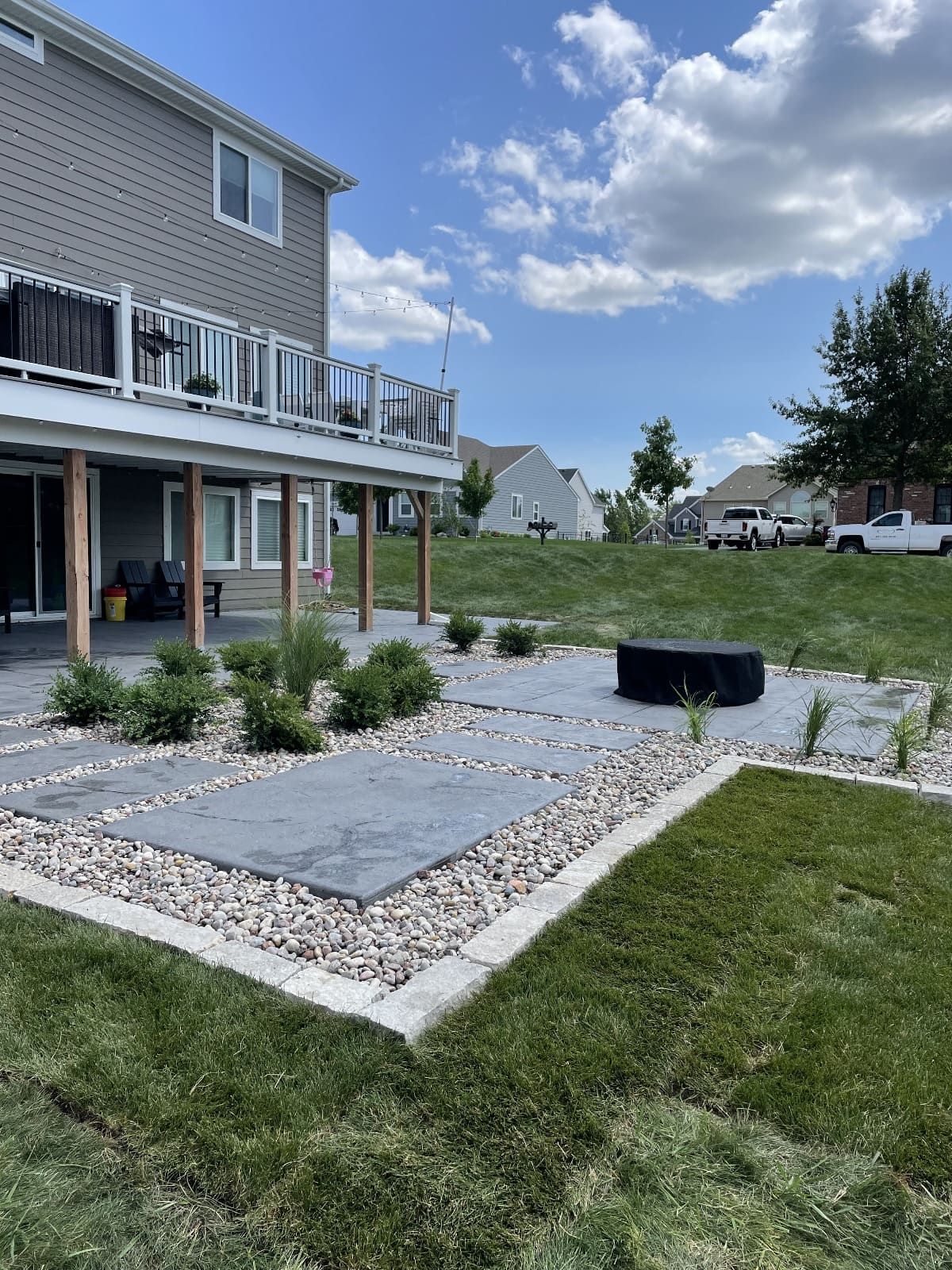 Backyard patio with gray pavers, gravel borders, and fire pit under a deck. Green grass and blue sky.