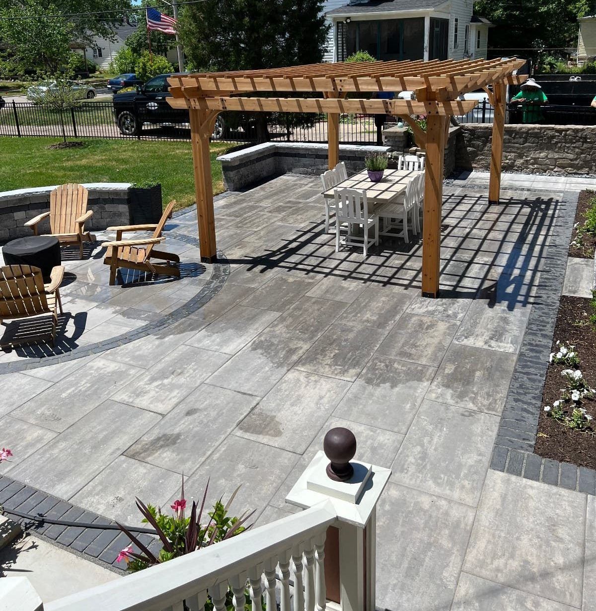 Patio with pergola, dining set, and fire pit on stone pavers, with a railing and green lawn.