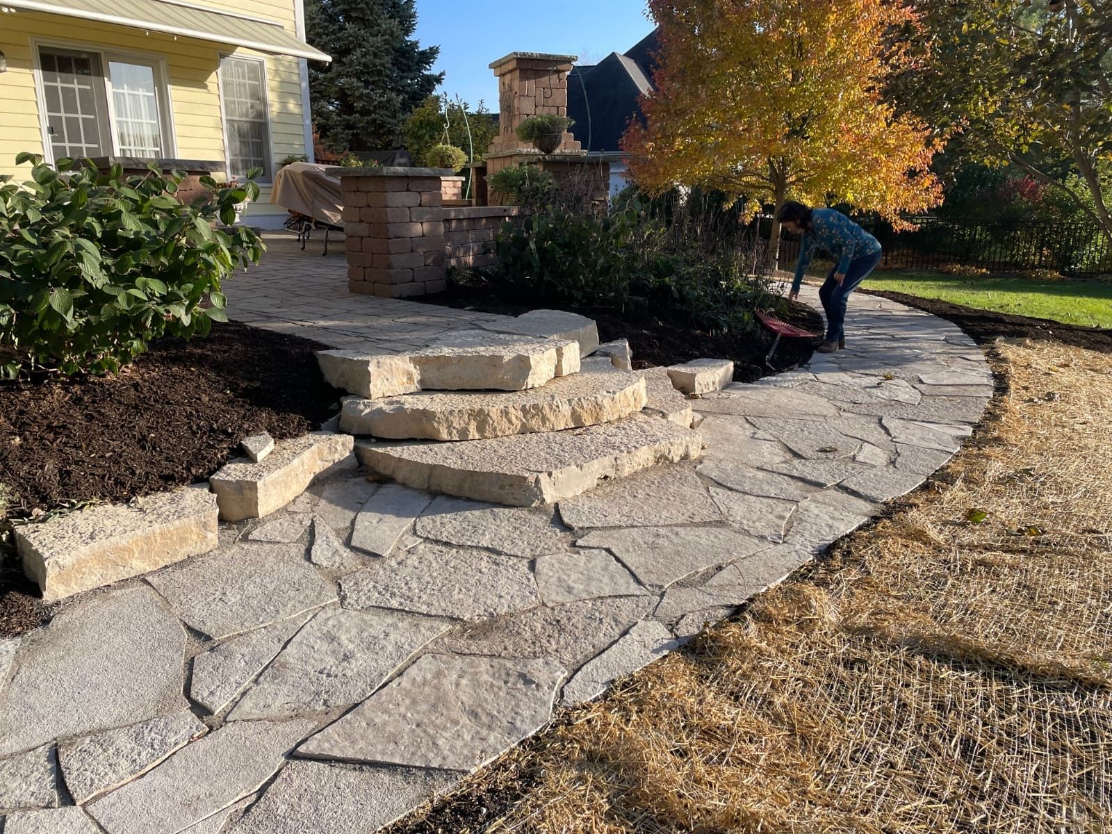 Stone pathway with steps, leading to a house. A person is tending to plants. Fall colors.