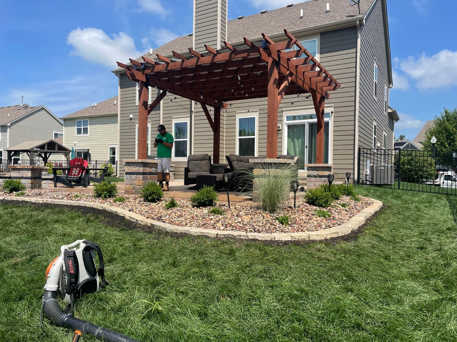 Backyard patio with pergola, landscaping, and a person using a leaf blower on a sunny day.