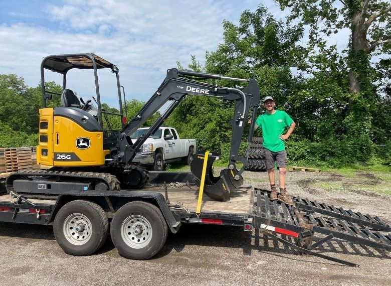 Man stands next to a John Deere mini-excavator on a trailer. Green shirt, sunny day.