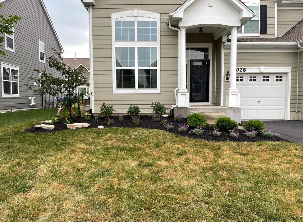 Tan house with a white garage door, front porch, and landscaped garden with dark mulch and various green plants.