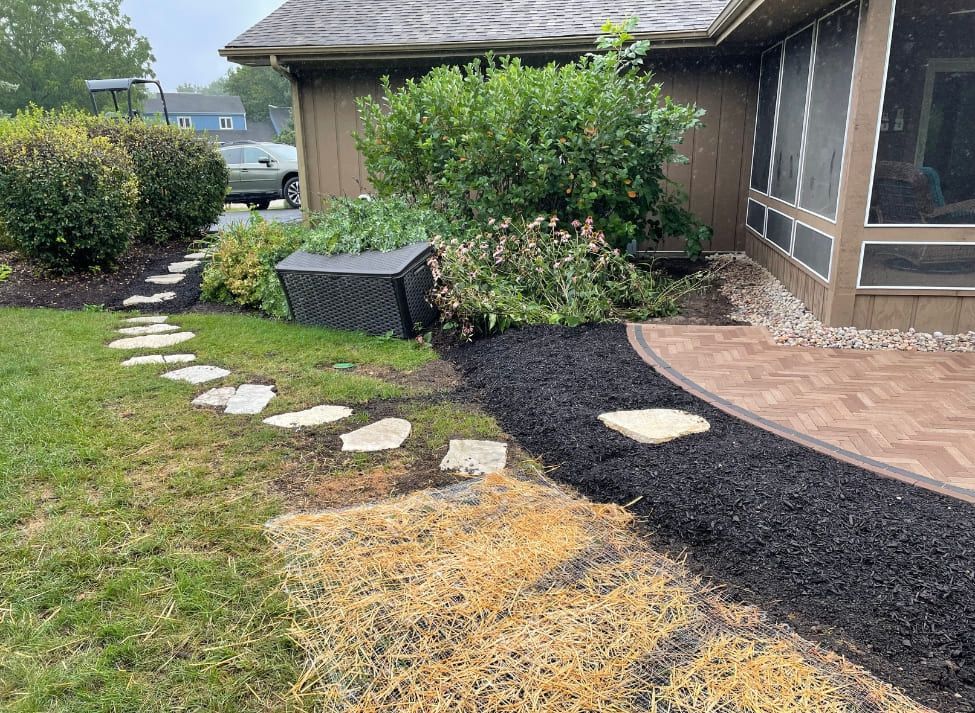 A stone path leads from grass to a brick patio next to a house with black mulch and bushes.