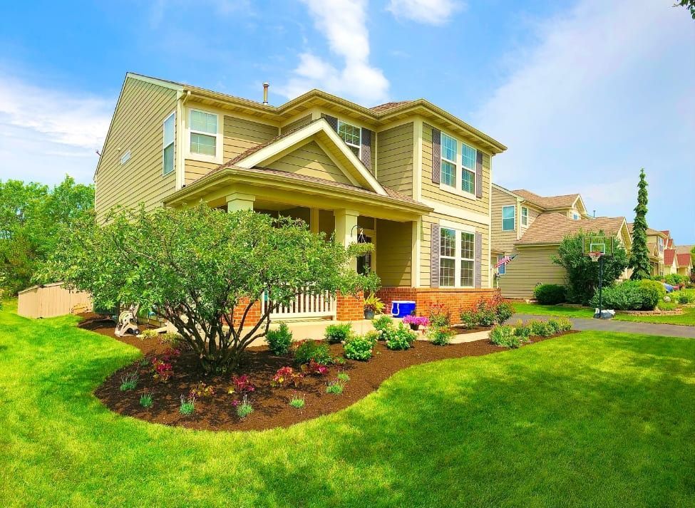 Two-story beige house with a porch and landscaped yard on a sunny day.