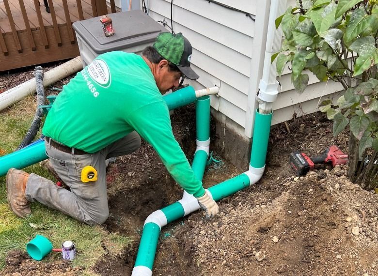 Person installing drainage pipes near a building. Green pipes and shirt, outdoor setting.