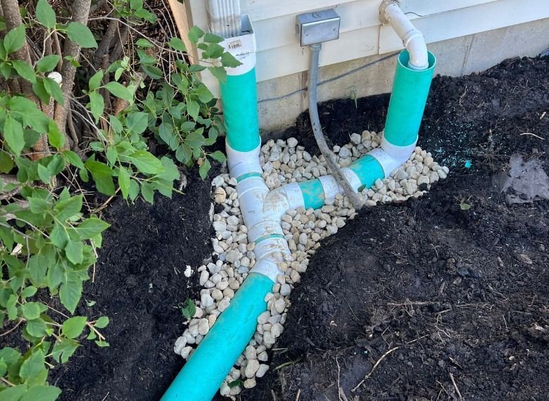 Green drainage pipes with gravel surround near a building, in a garden bed.