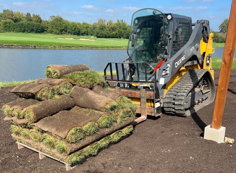 John Deere skid steer carrying pallets of rolled turf by a lake.