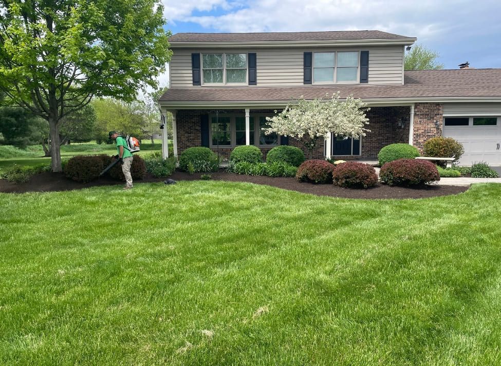 Person mulching garden beds in front of a two-story house with green lawn.
