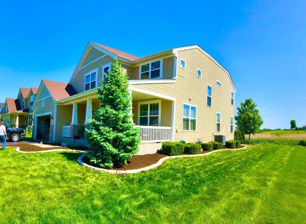 Two-story tan house with a green lawn, blue sky, and a tall evergreen tree in front.