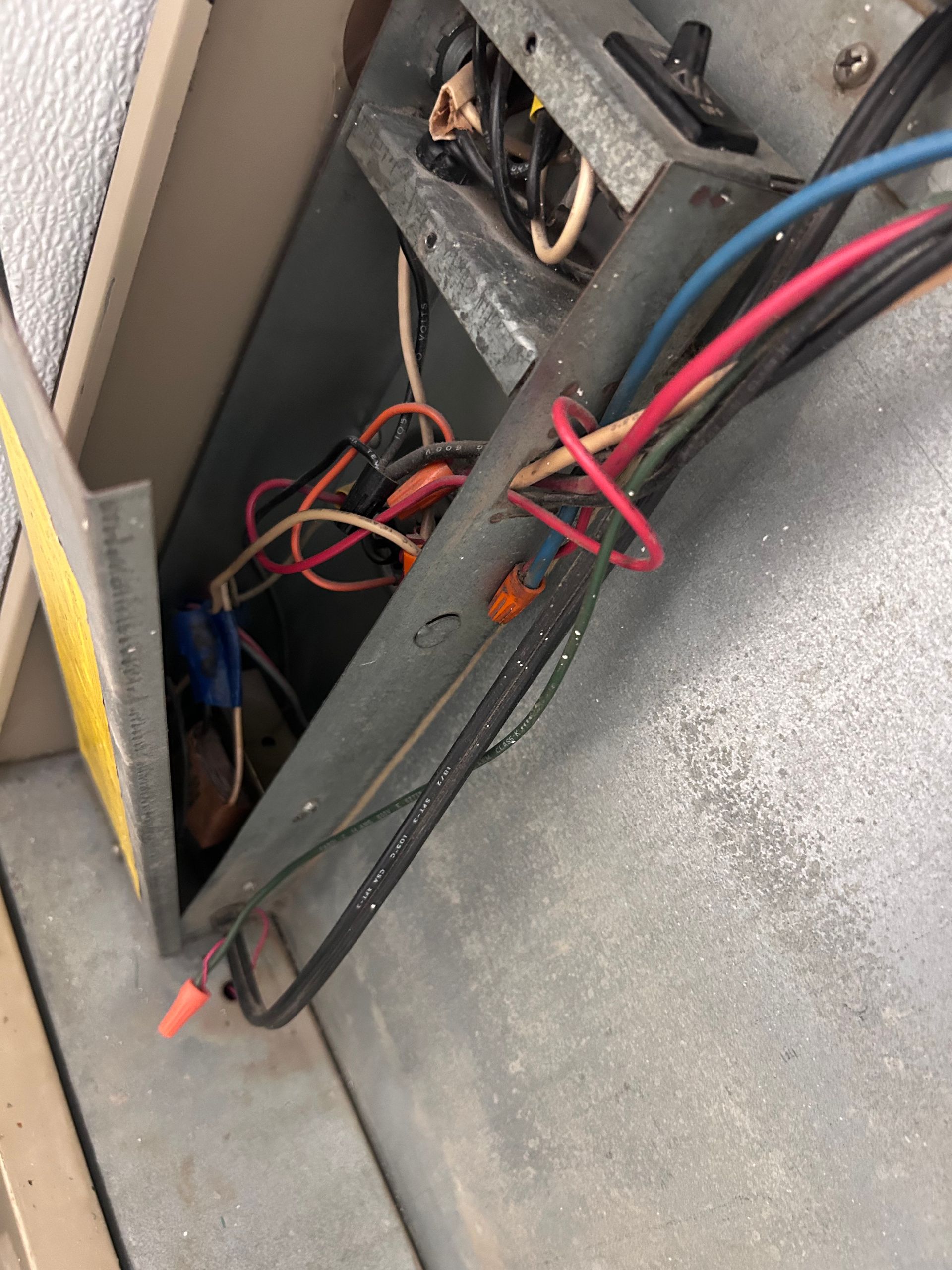 A close-up of exposed, disorganized electrical wiring and wire nuts inside an open metal housing panel of an HVAC unit.