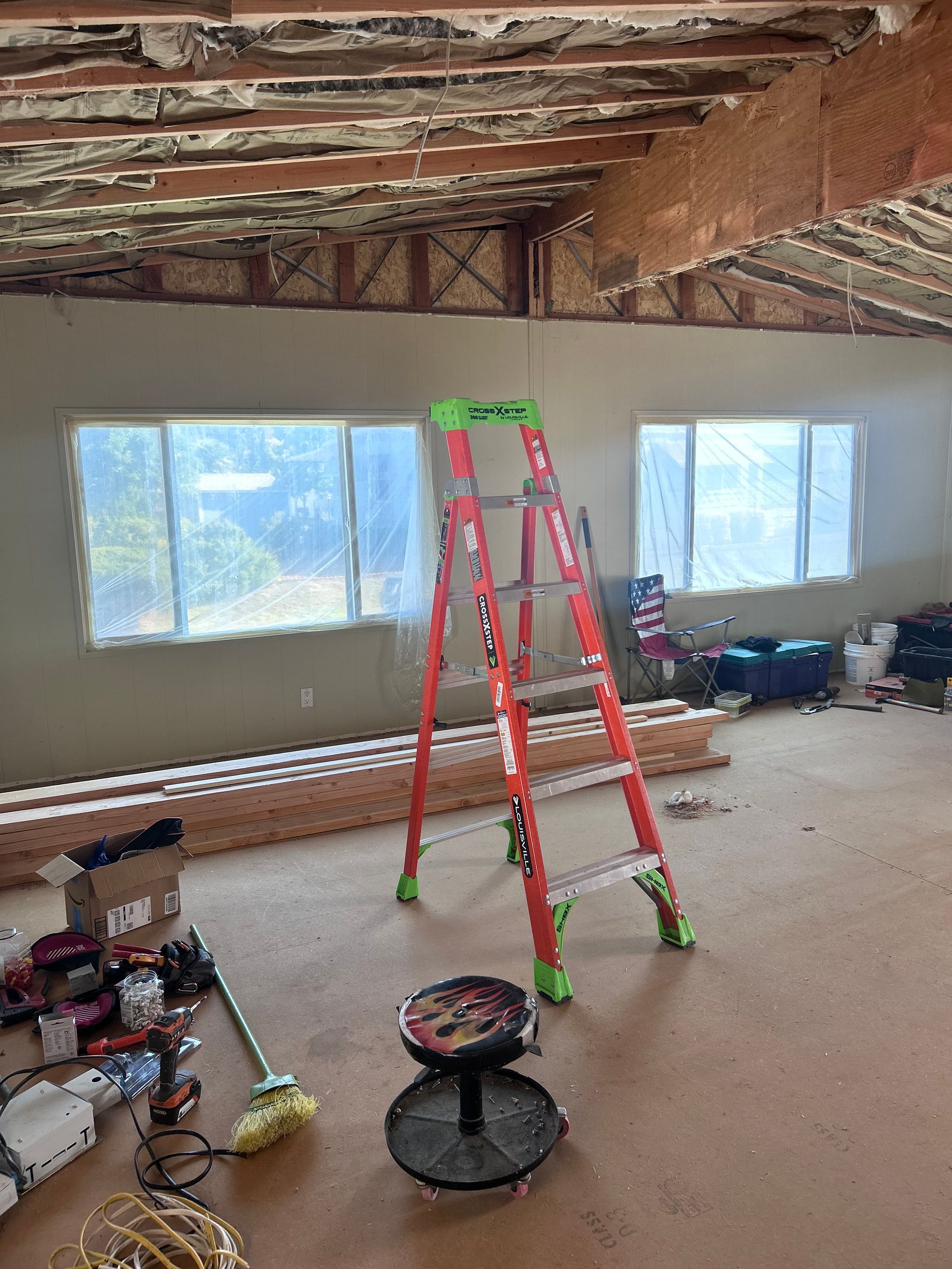 A construction site in a room with a red ladder, a rolling stool, tools on the floor, and an exposed ceiling structure.