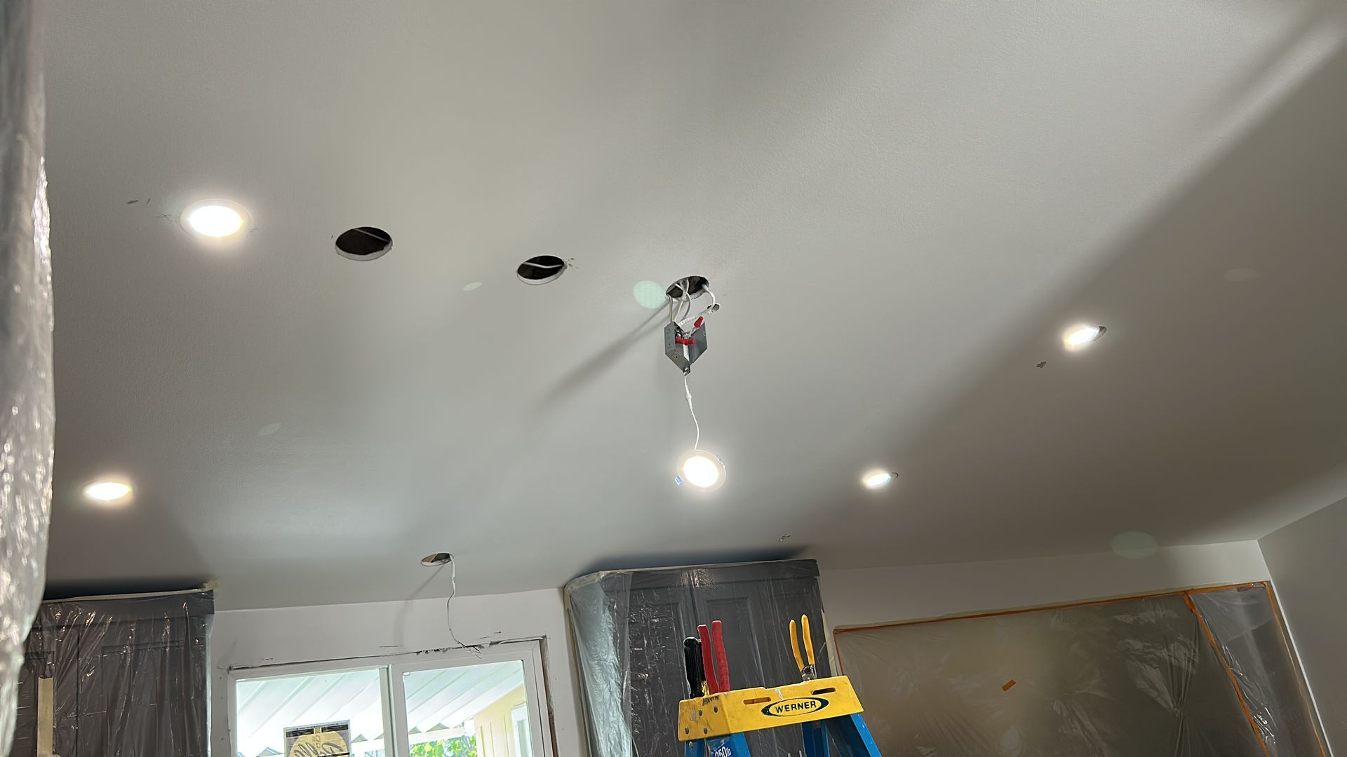 White ceiling under construction with several recessed lights, empty light fixture holes, and a yellow stepladder below.