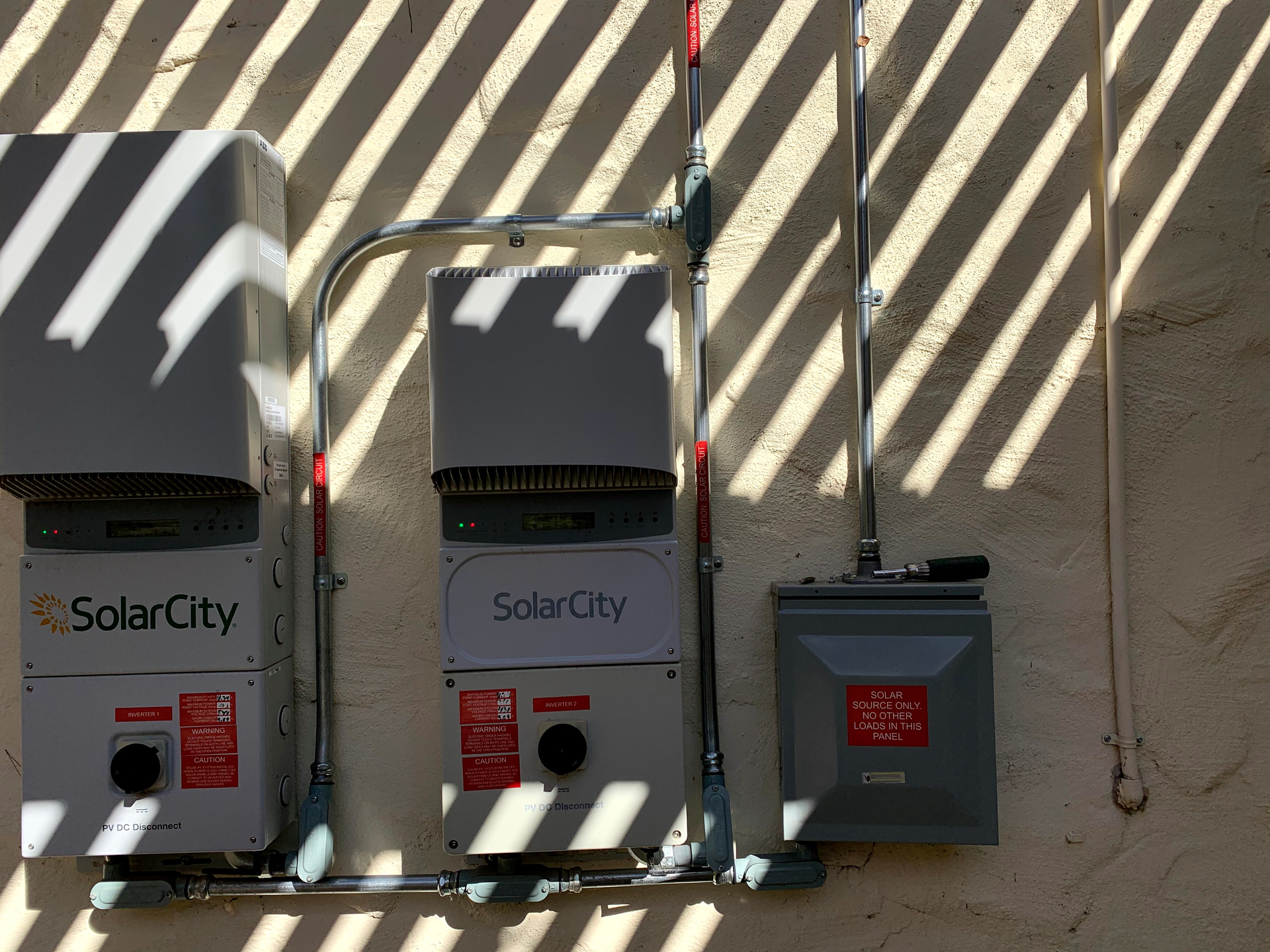 Two SolarCity inverters and a grey electrical disconnect box mounted on an exterior wall with shadow-striped lighting.