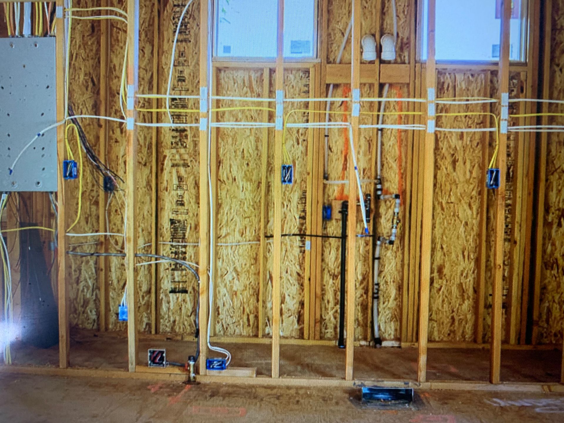 Framed interior wall of a house under construction with exposed electrical wiring, plumbing pipes, and electrical boxes.