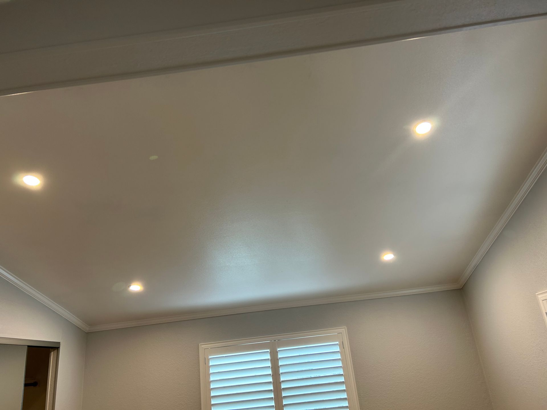 A white room ceiling with crown molding and four recessed lights, featuring a window with white plantation shutters.