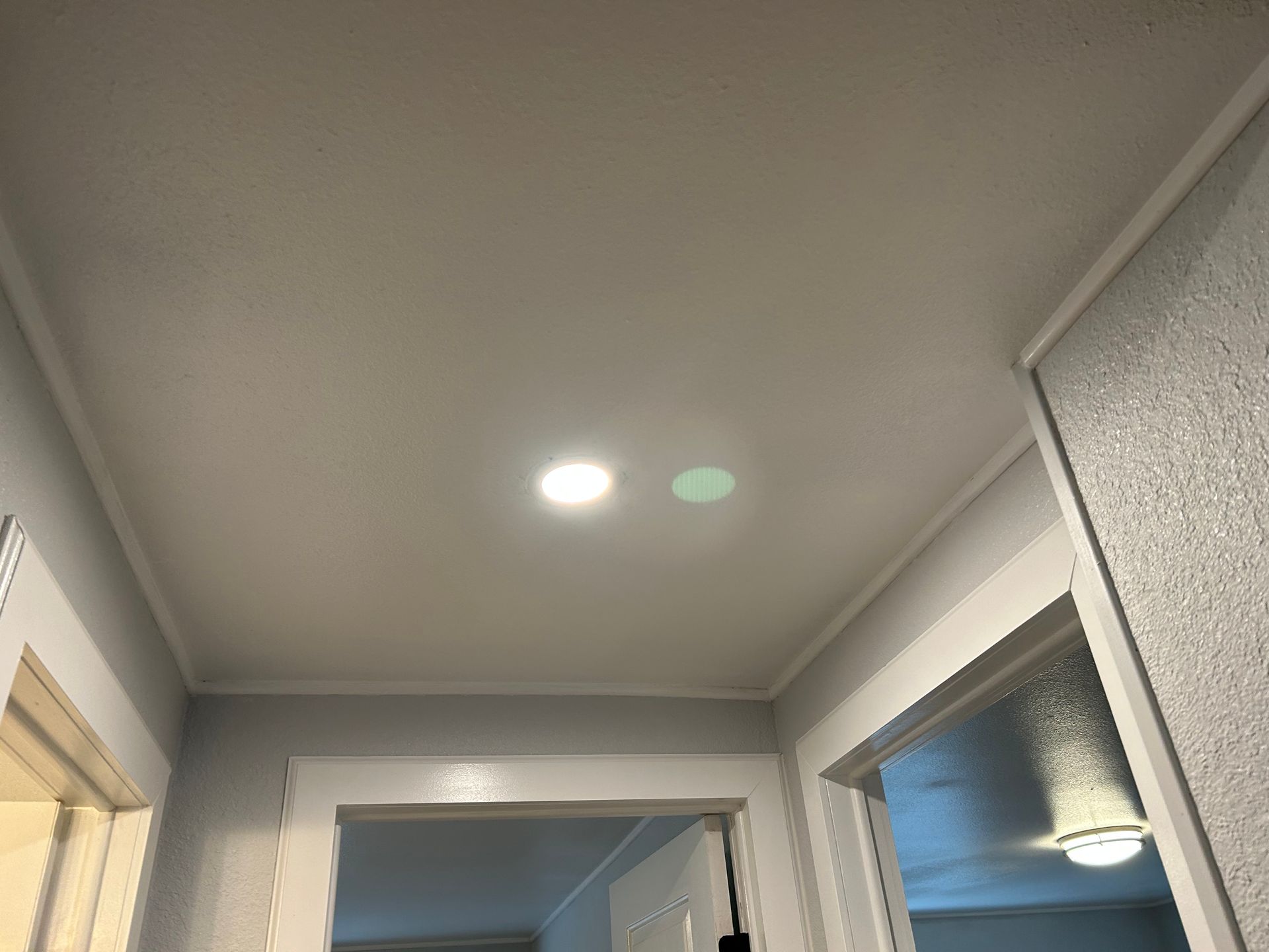 A hallway view showing a white ceiling with a lit recessed light and a green lens flare, flanked by doorways.