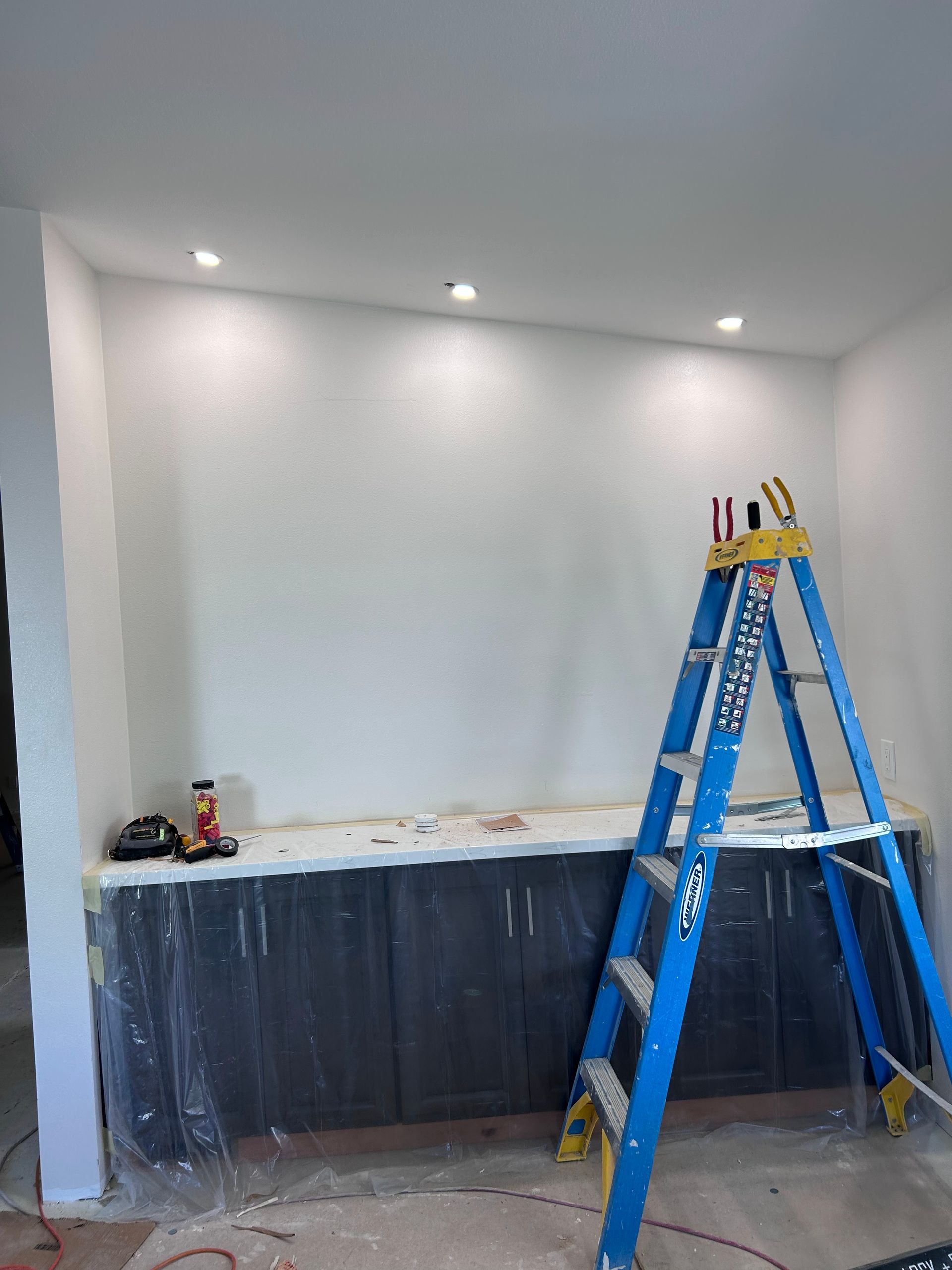 A blue step ladder stands in front of a wall with newly installed dark cabinets and three recessed ceiling lights.