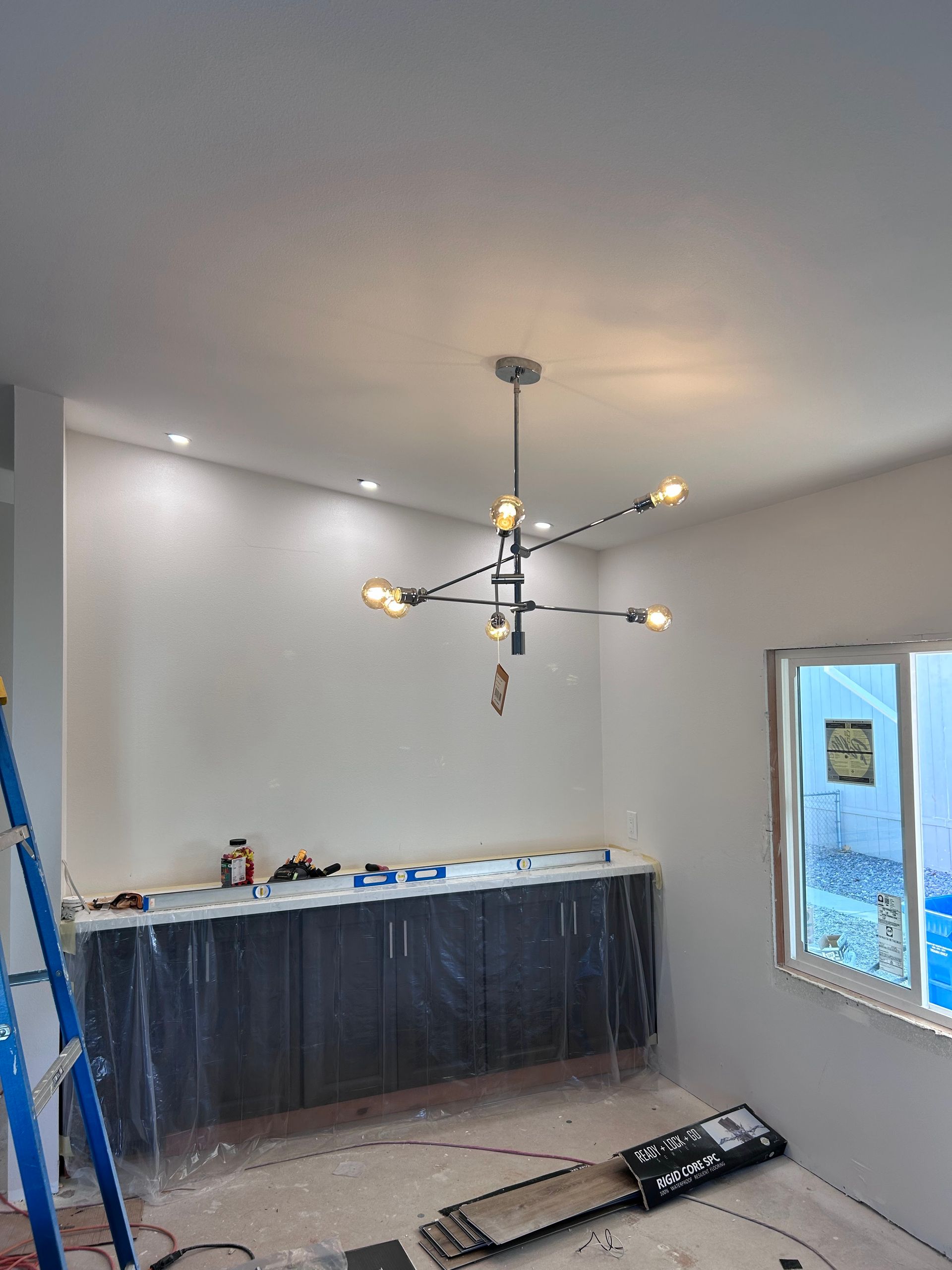 A modern black chandelier hangs above a dark, plastic-covered cabinet in a room undergoing renovation.