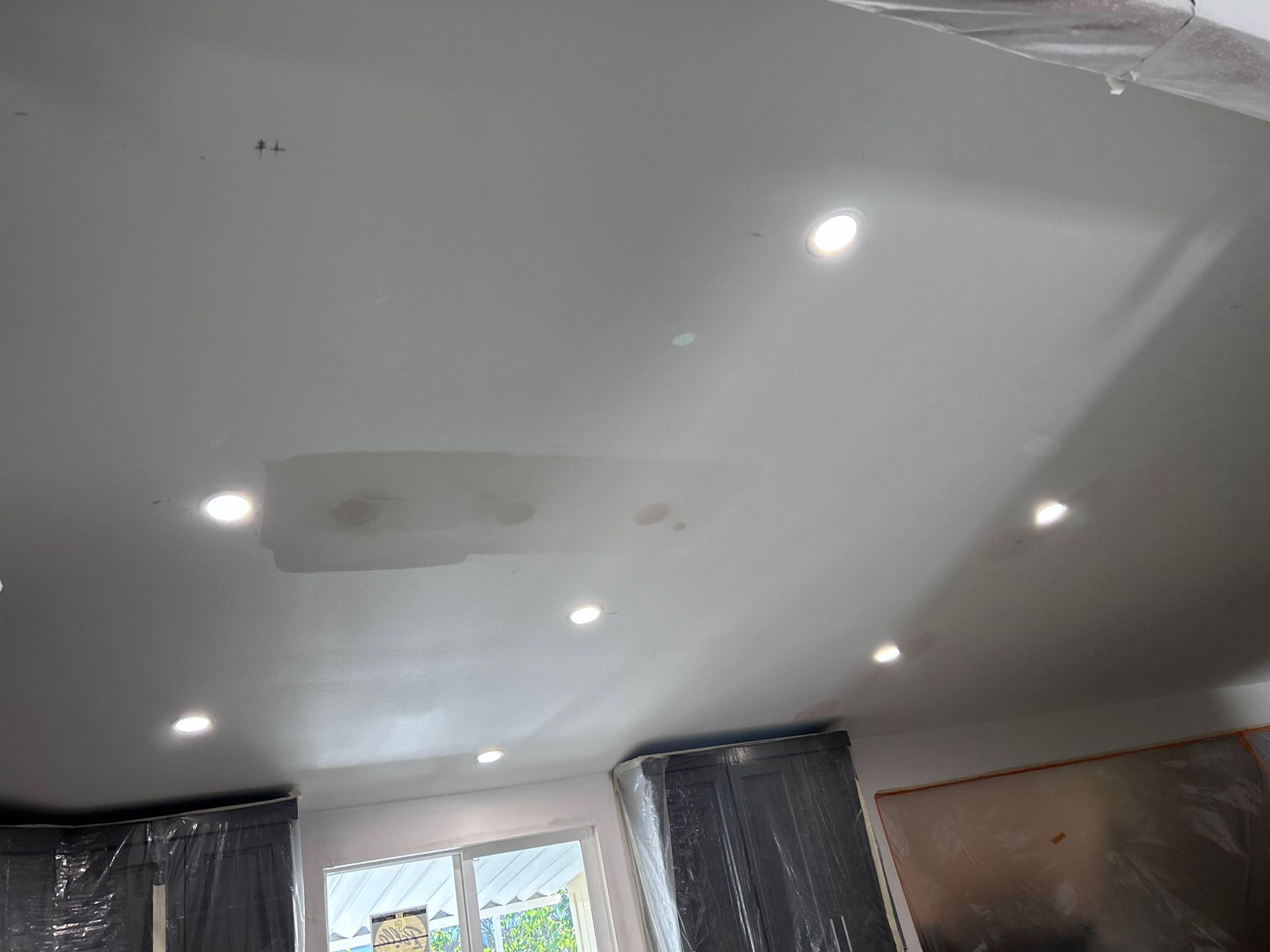 A ceiling with multiple recessed lights and a discolored patch in the center, partially covered with plastic sheeting.