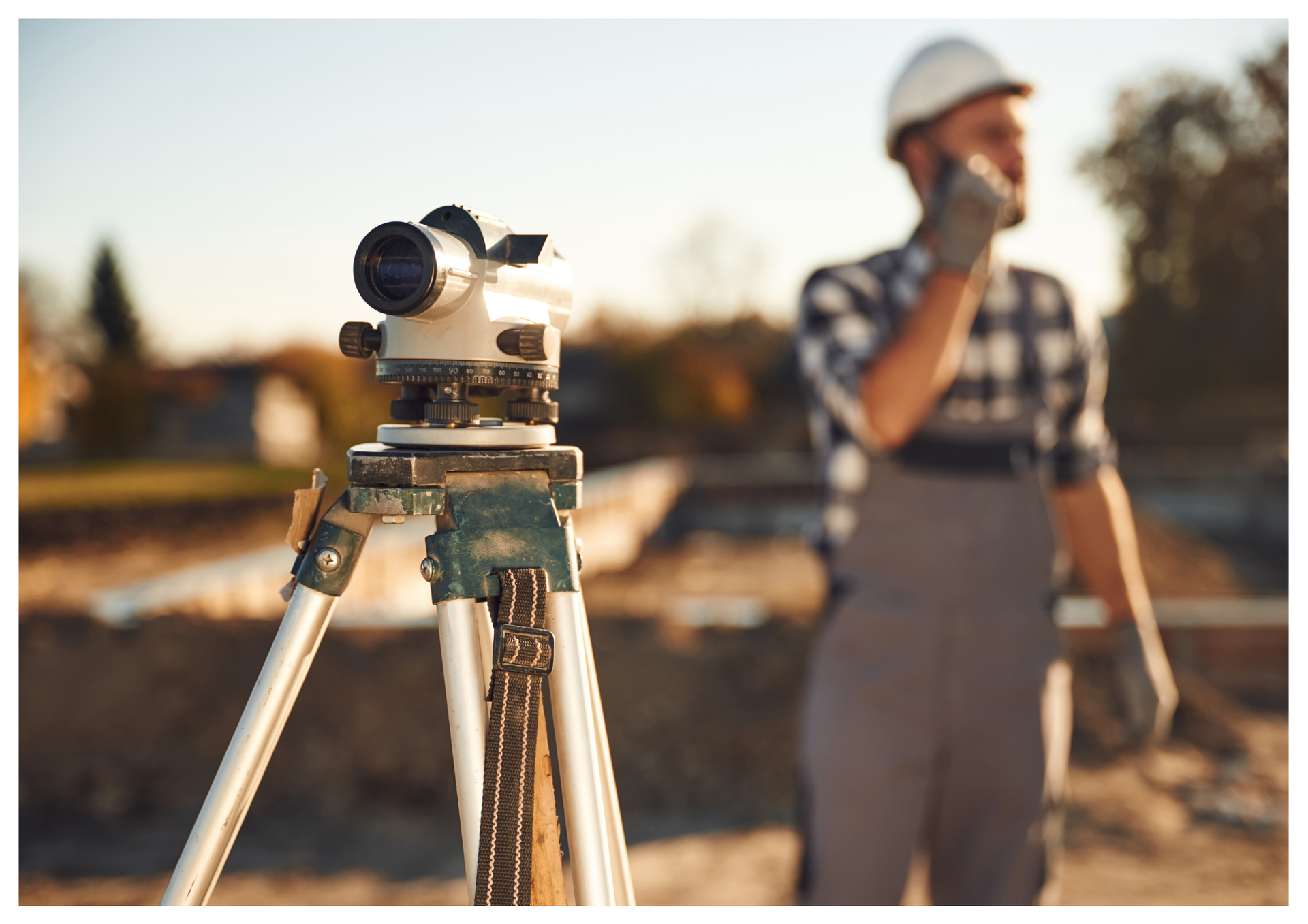 Ant trikojo pastatyta matininko įranga, fone – matininkas, kalbantis telefonu.