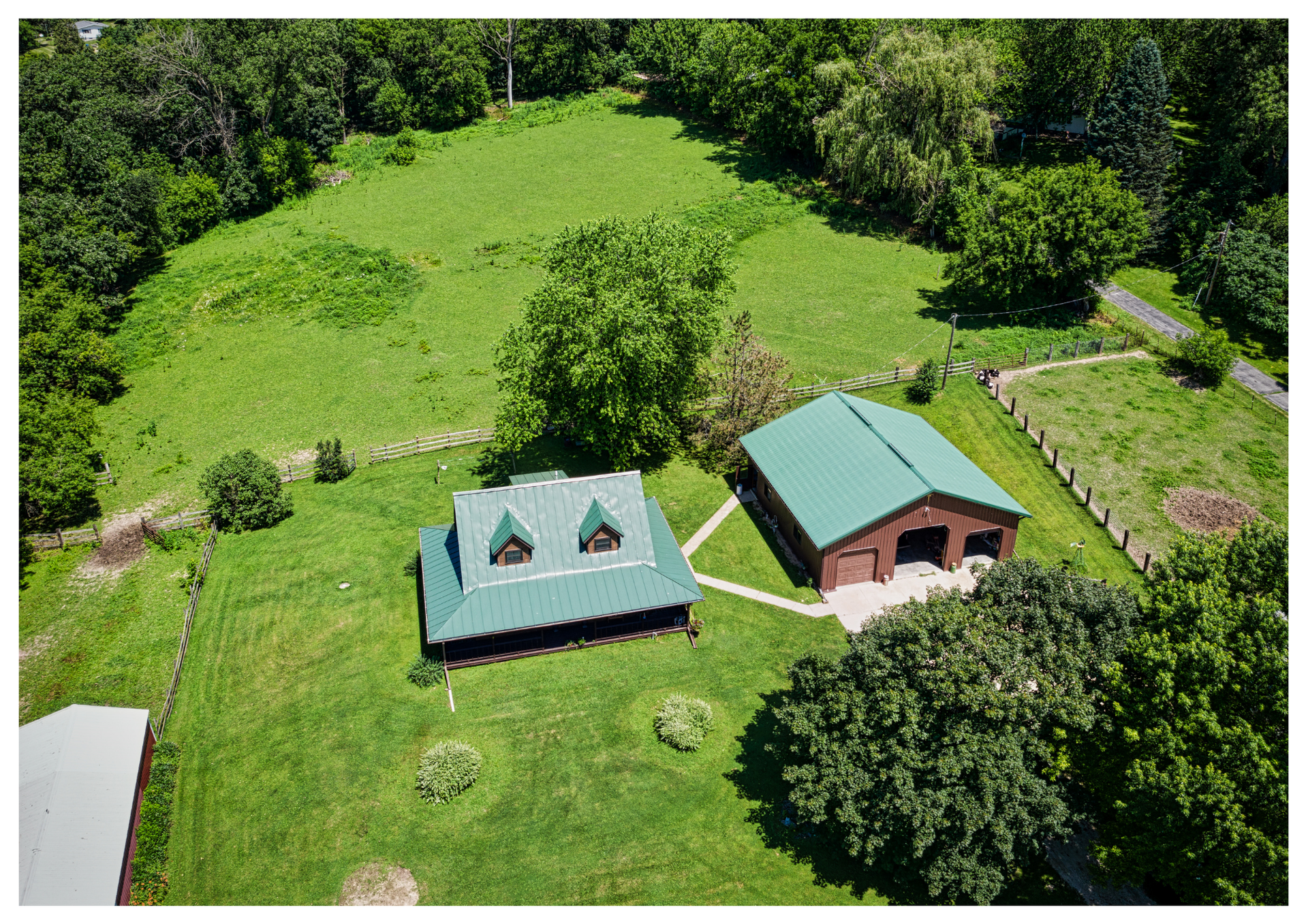 Namas ir garažas su žaliais stogais iš oro, apsupti žolėto kiemo ir medžių.