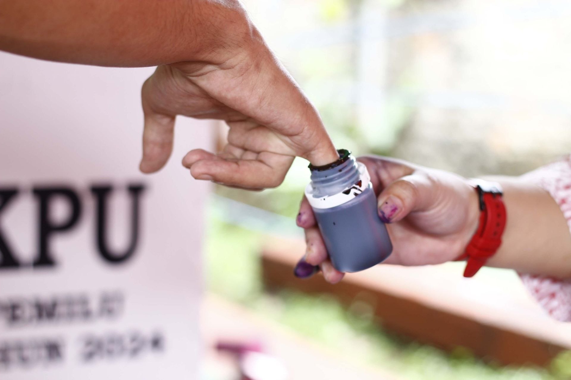 A person is putting purple ink into another person 's finger in front of a sign that says kpu