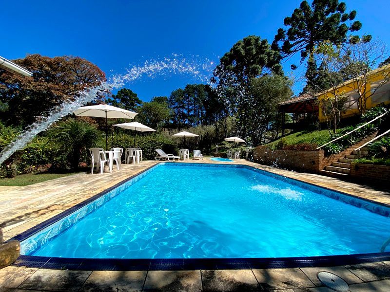 Piscina azul com fonte, guarda-sóis brancos e cadeiras. Árvores verdejantes sob um céu azul brilhante.