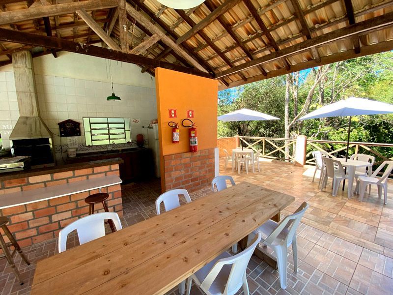 Cozinha externa coberta com mesa comprida de madeira, detalhes em tijolo aparente e cadeiras de plástico brancas. Guarda-sóis e árvores visíveis.