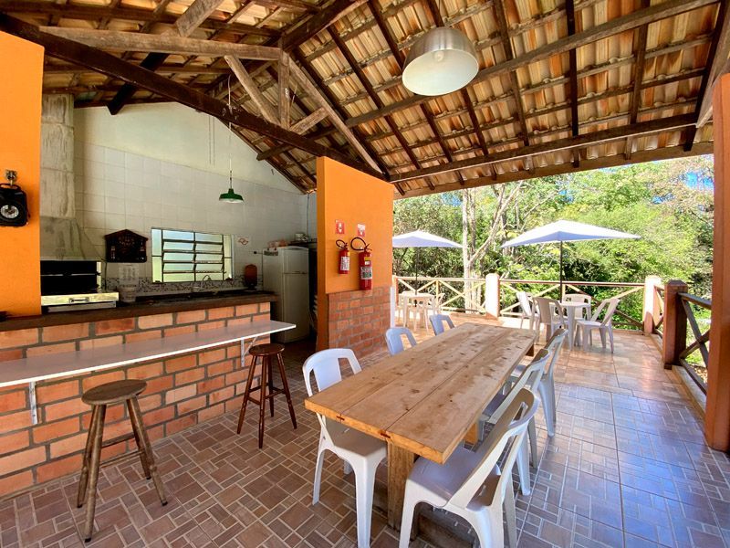 Cozinha e área de jantar externas com detalhes em tijolo aparente, mesa e cadeiras de madeira e vegetação ao redor.
