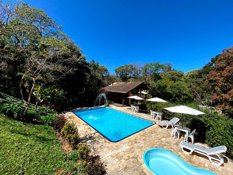 Área da piscina com água turquesa rodeada de árvores, com espreguiçadeiras e guarda-sóis sob um céu azul e límpido.