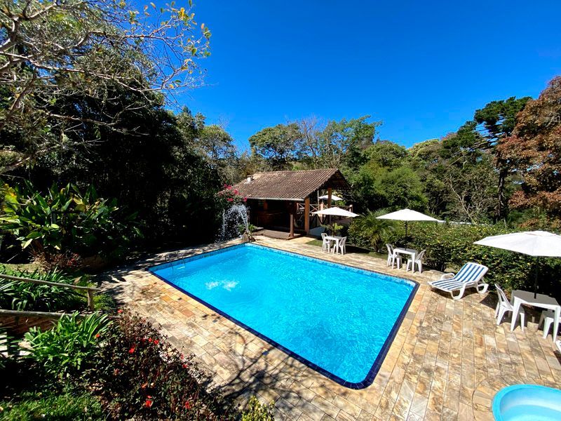 Piscina rodeada por um pátio de pedra, árvores, uma pequena construção e mesas ao ar livre com guarda-sóis. Céu azul.