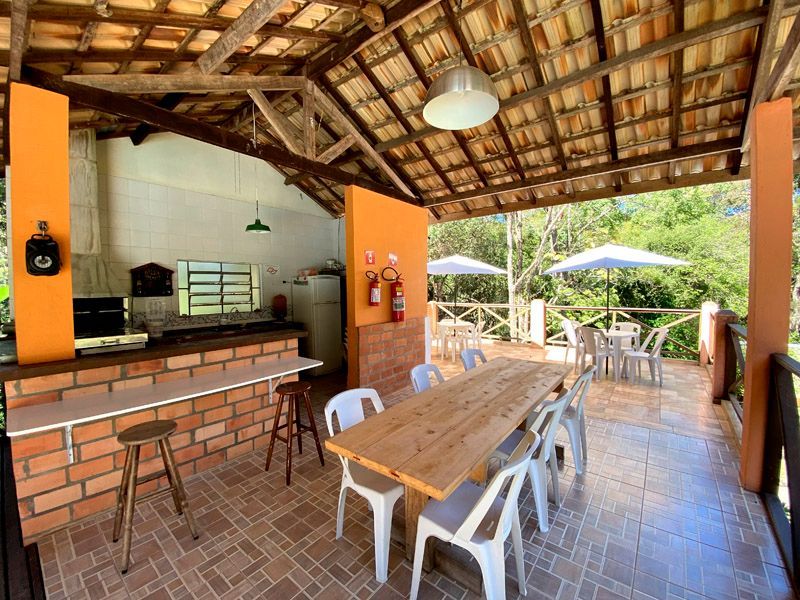 Cozinha externa com uma longa mesa e cadeiras de madeira, bancada de tijolos e detalhes em laranja.