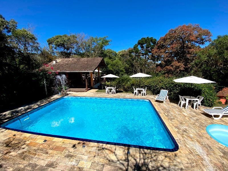 Piscina com água azul e pátio ao redor com mesas, cadeiras e guarda-sóis. Prédio e árvores ao fundo.
