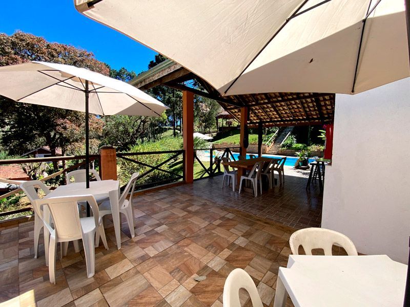 Pátio com mesas brancas e guarda-sóis com vista para uma piscina e árvores em um dia ensolarado.