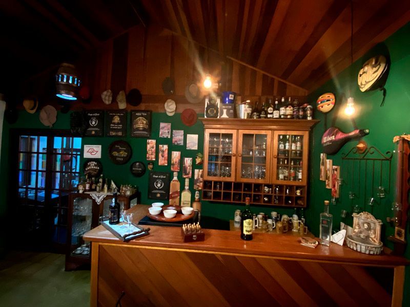 Interior de bar aconchegante com balcão de madeira, paredes verdes, garrafas de bebidas alcoólicas e itens decorativos.