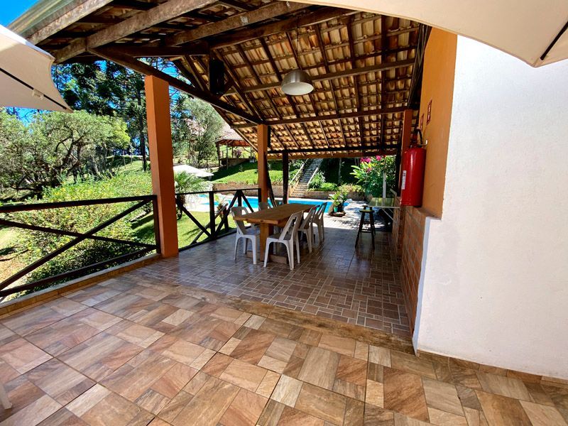 Varanda coberta com mesa de jantar e vista para a piscina. Piso de azulejo marrom, vigas de madeira, árvores e céu azul.