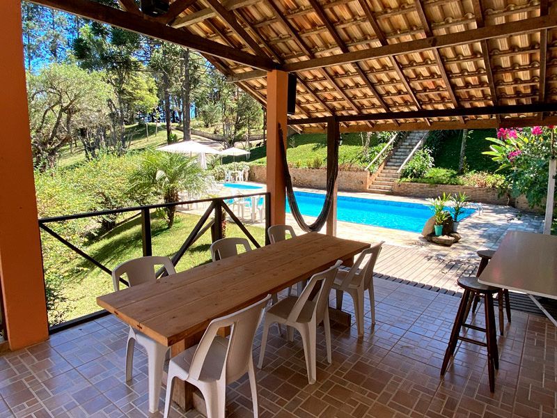 Pátio coberto com uma longa mesa de madeira e cadeiras com vista para uma piscina e vegetação exuberante.